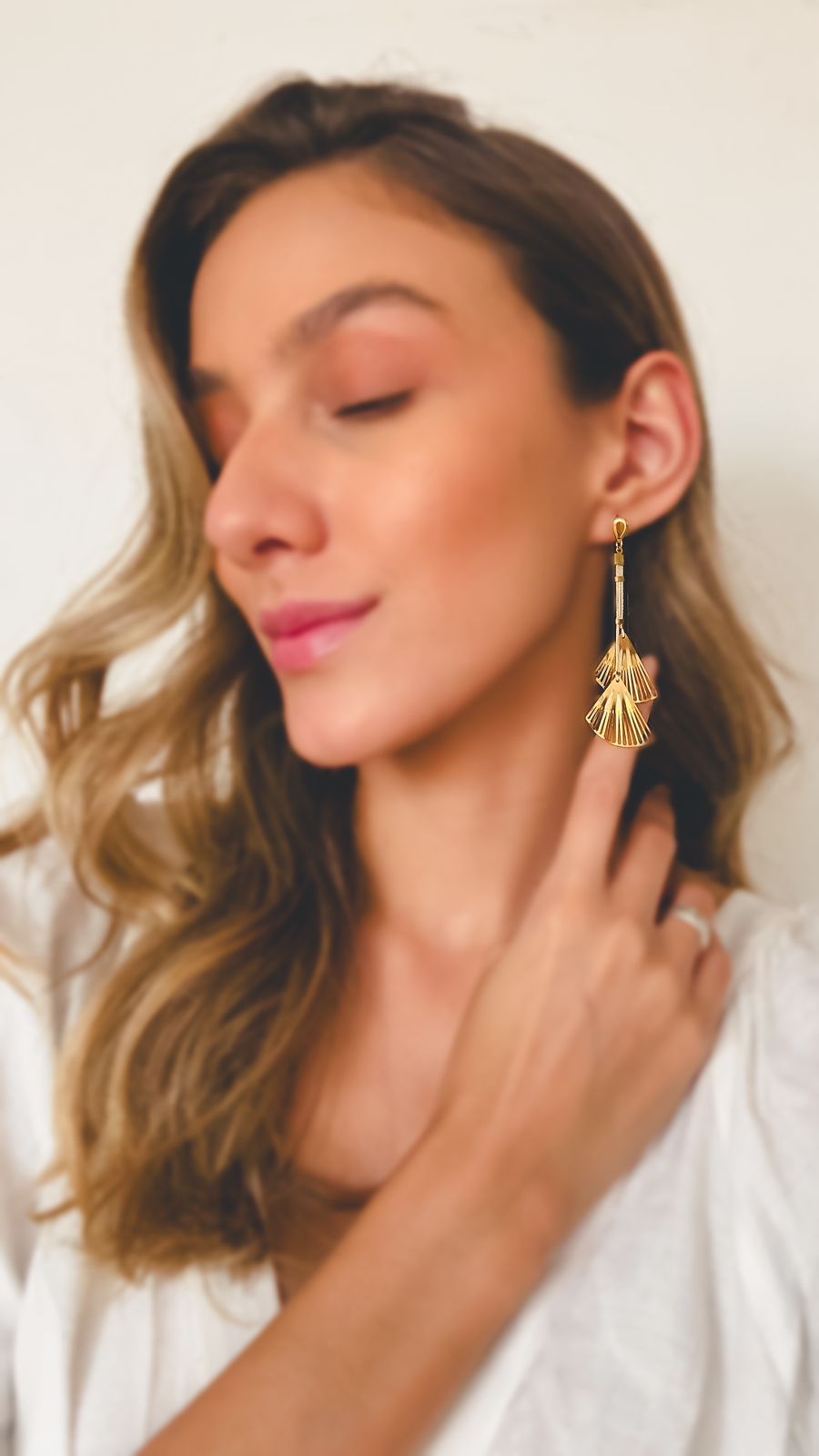 Mujer feliz usando Pendientes largos de Purplêe Store hechos a mano en palha de burití con colgantes dorados en forma de abanico – joyas sostenibles y sofisticadas