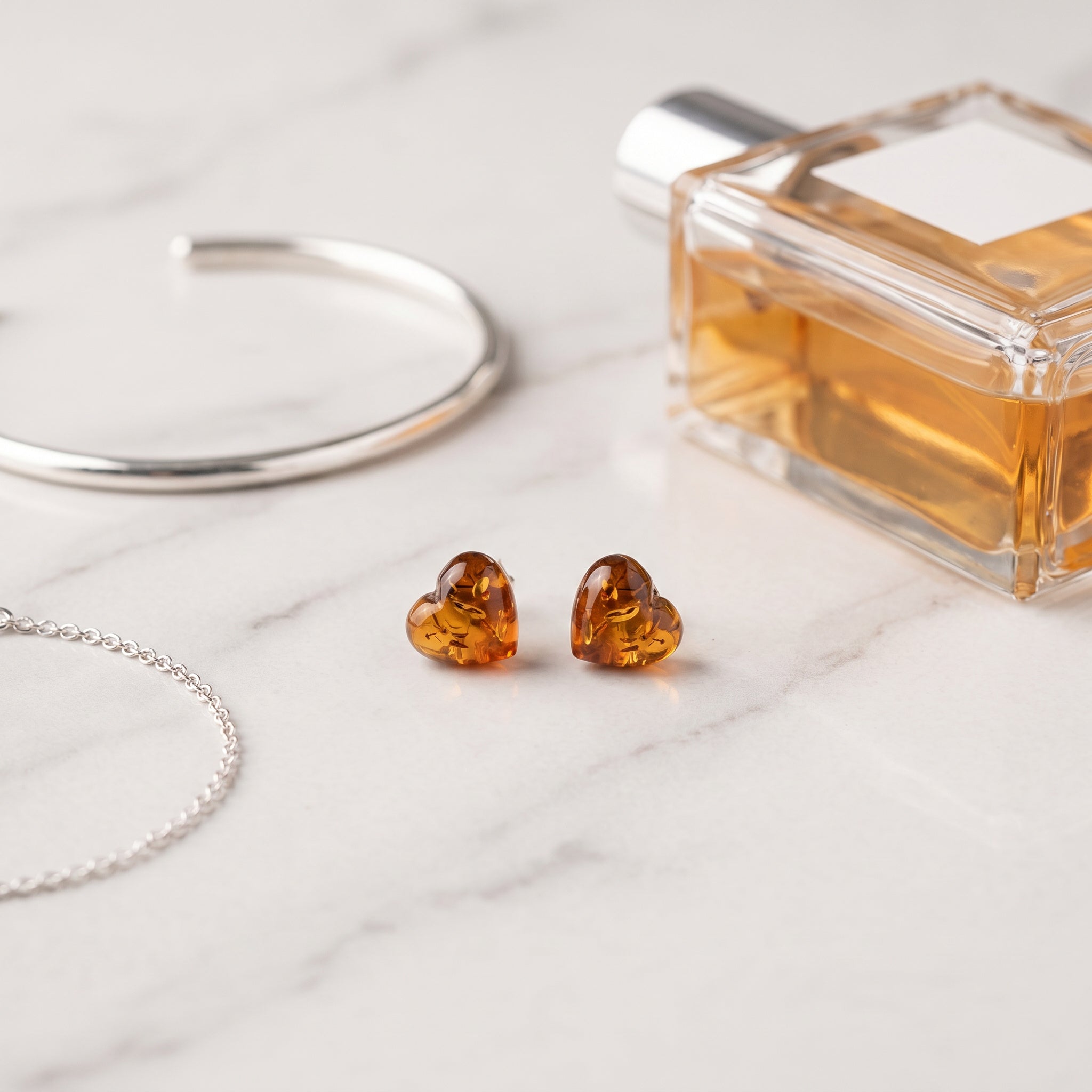 Amber Heart 925 Silver Studs|Purplee Store next to a perfume bottle and silver bracelet on a light surface.